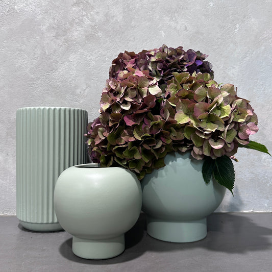 Three vases with distinct shapes are displayed together. Two belong to the Sage Ceramic Vaseware collection by Flowers on Norton St and are empty, while the other contains a cluster of wilted hydrangea flowers in green and purple tones, set against a textured gray background.