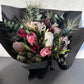 Introducing the "Little Native Bunch" by Flowers on Norton St: A striking bouquet showcasing native flora and a diverse array of protea flowers, complemented with lush greenery and subtle pink accents. This exquisite arrangement is beautifully wrapped in elegant black paper and tied with a chic black ribbon, all set against a light gray backdrop.