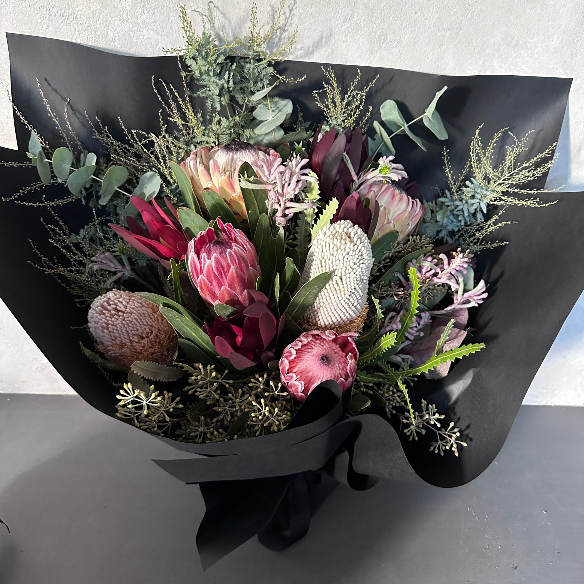 Introducing the "Little Native Bunch" by Flowers on Norton St: A striking bouquet showcasing native flora and a diverse array of protea flowers, complemented with lush greenery and subtle pink accents. This exquisite arrangement is beautifully wrapped in elegant black paper and tied with a chic black ribbon, all set against a light gray backdrop.