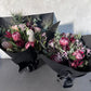 The Little Native Bunch 2024 by Flowers on Norton St features two statement bouquets of native Australian flora, with proteas and banksias wrapped in black paper, enhanced by dark ribbons. Seasonal pink and cream flowers are complemented by green foliage on a neutral background.