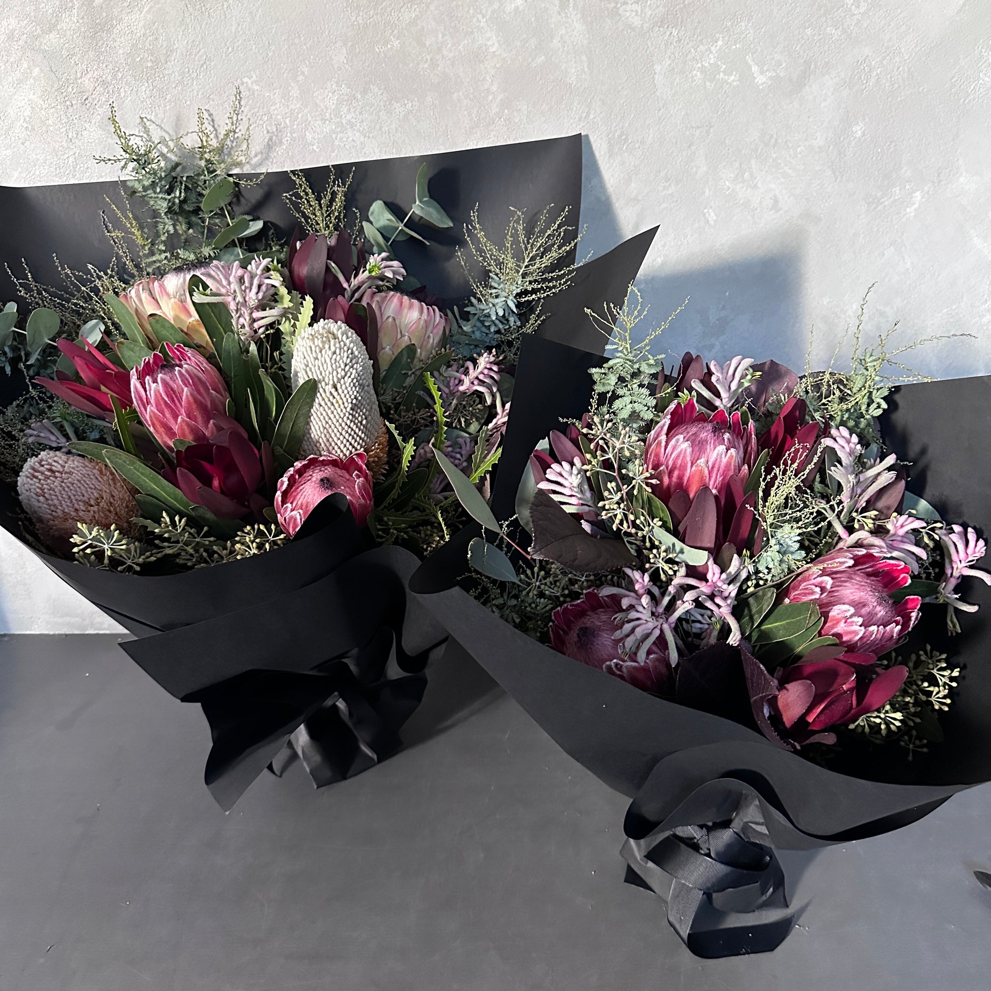 The Little Native Bunch 2024 by Flowers on Norton St features two statement bouquets of native Australian flora, with proteas and banksias wrapped in black paper, enhanced by dark ribbons. Seasonal pink and cream flowers are complemented by green foliage on a neutral background.