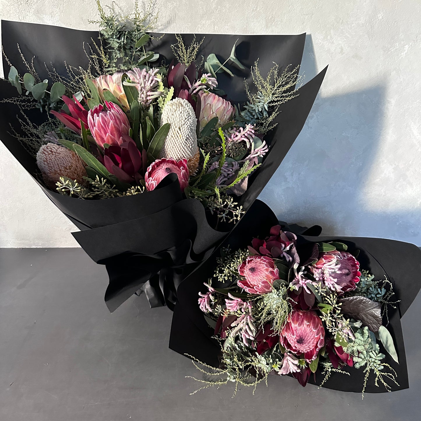 Displayed against a light gray background, two elegant bouquets from Flowers on Norton St's Little Native Bunch 2024 feature pink and white protea flowers accented with eucalyptus leaves and delicate greenery, all wrapped in black paper.