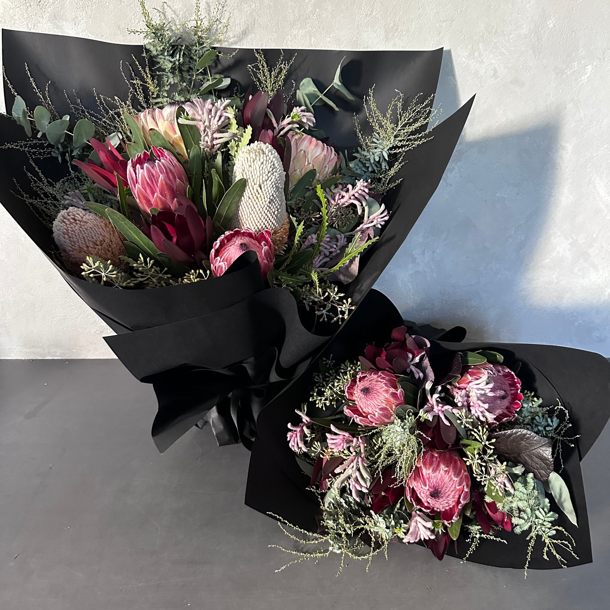 Displayed against a light gray background, two elegant bouquets from Flowers on Norton St's Little Native Bunch 2024 feature pink and white protea flowers accented with eucalyptus leaves and delicate greenery, all wrapped in black paper.