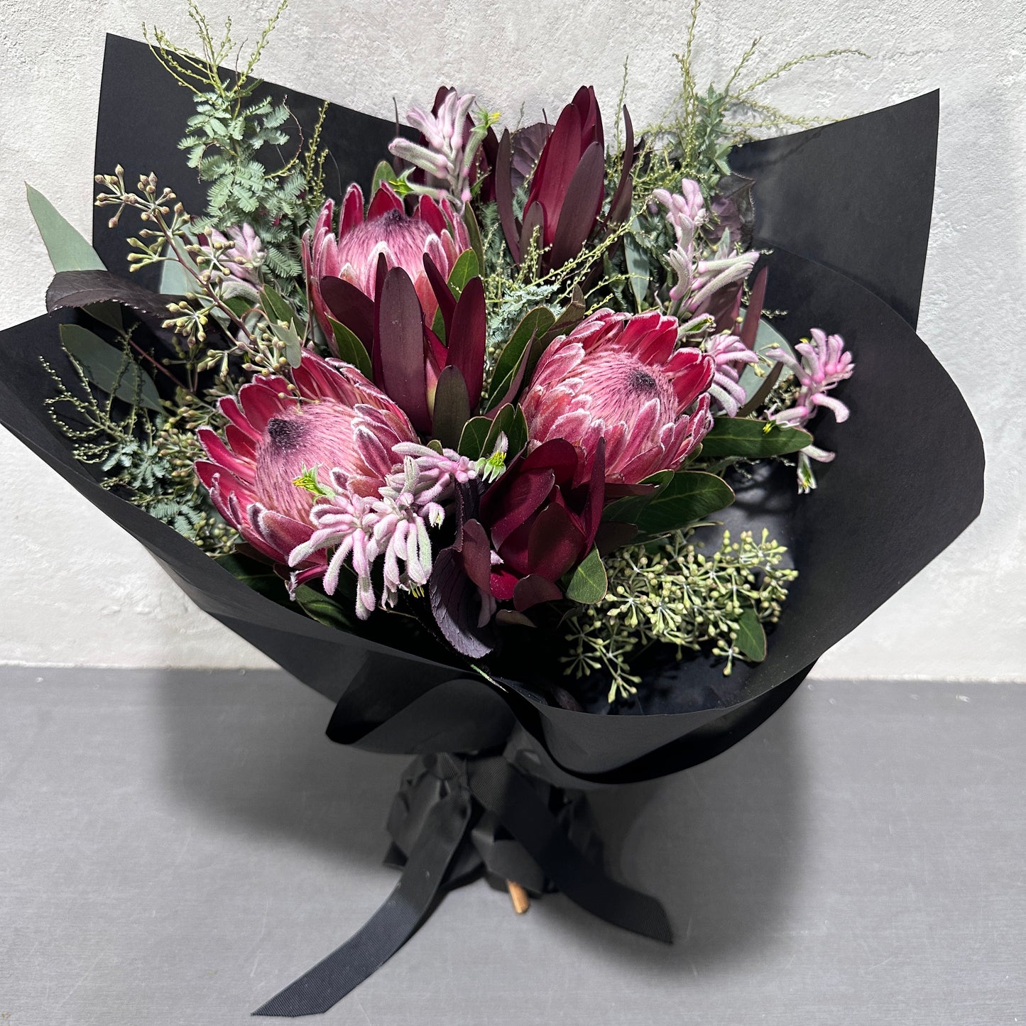 The "Little Native Bunch 2024" by Flowers on Norton St is a striking bouquet of pink protea, deep burgundy leaves, and delicate greenery, wrapped in black paper with a ribbon. This native flora arrangement is elegantly displayed on a gray surface against a light wall.