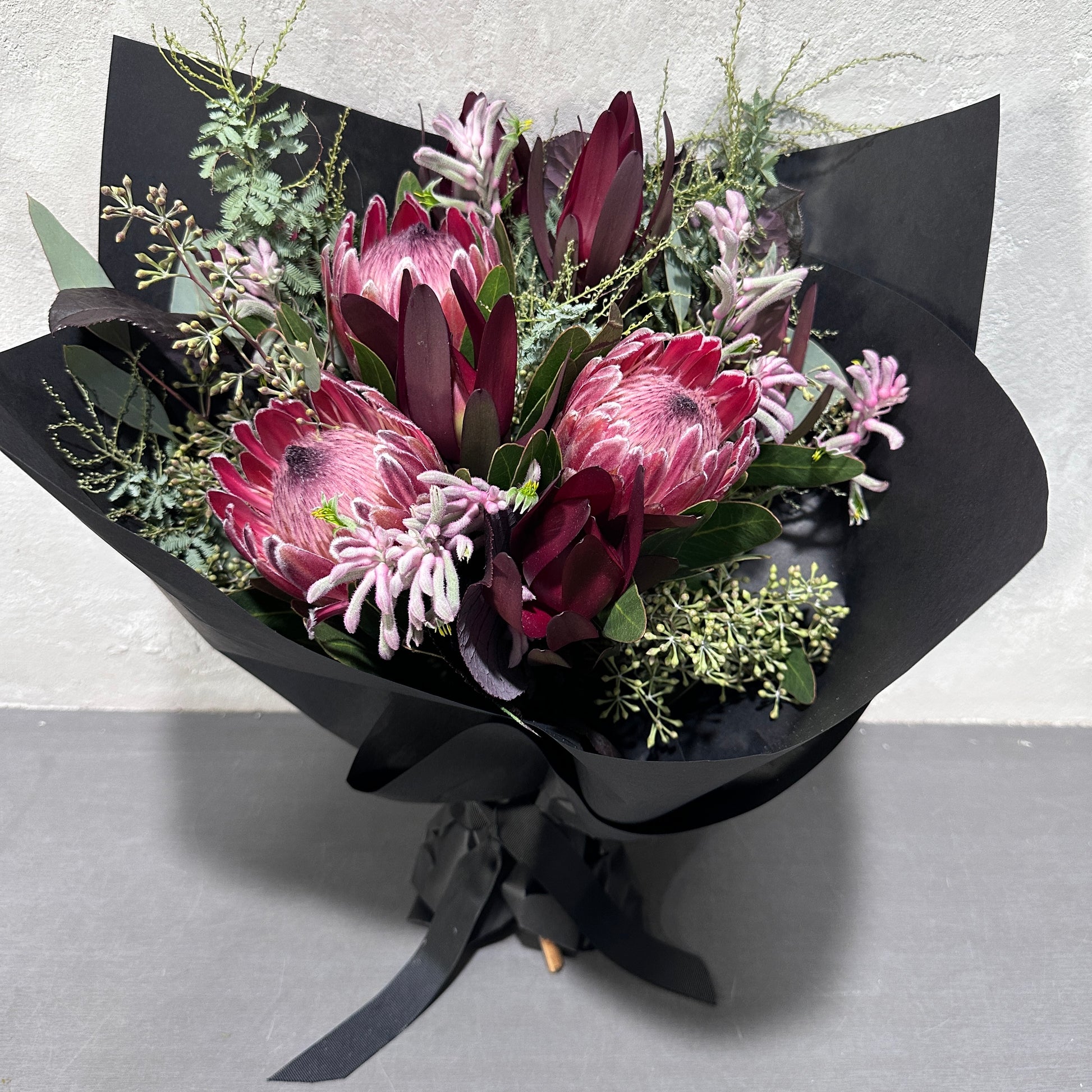 The "Little Native Bunch 2024" by Flowers on Norton St is a striking bouquet of pink protea, deep burgundy leaves, and delicate greenery, wrapped in black paper with a ribbon. This native flora arrangement is elegantly displayed on a gray surface against a light wall.