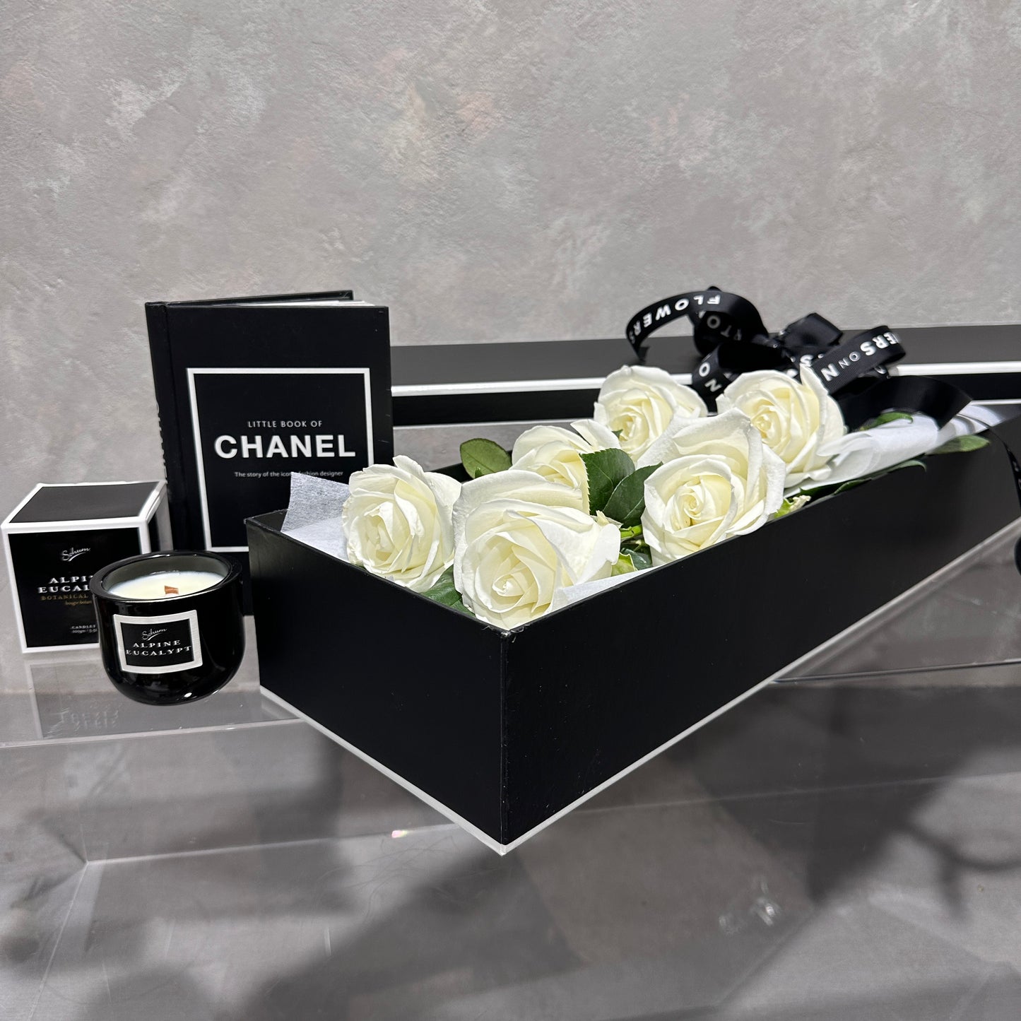 A FlowersonNortonSt Rose Gift Box, featuring a sophisticated black exterior and filled with pristine white roses, is elegantly displayed on a clear surface. Nearby, you can find a stylish black candle in a jar and the chic "Little Book of Chanel" in black. A striking black ribbon with white text adds to this memorable gift setting.