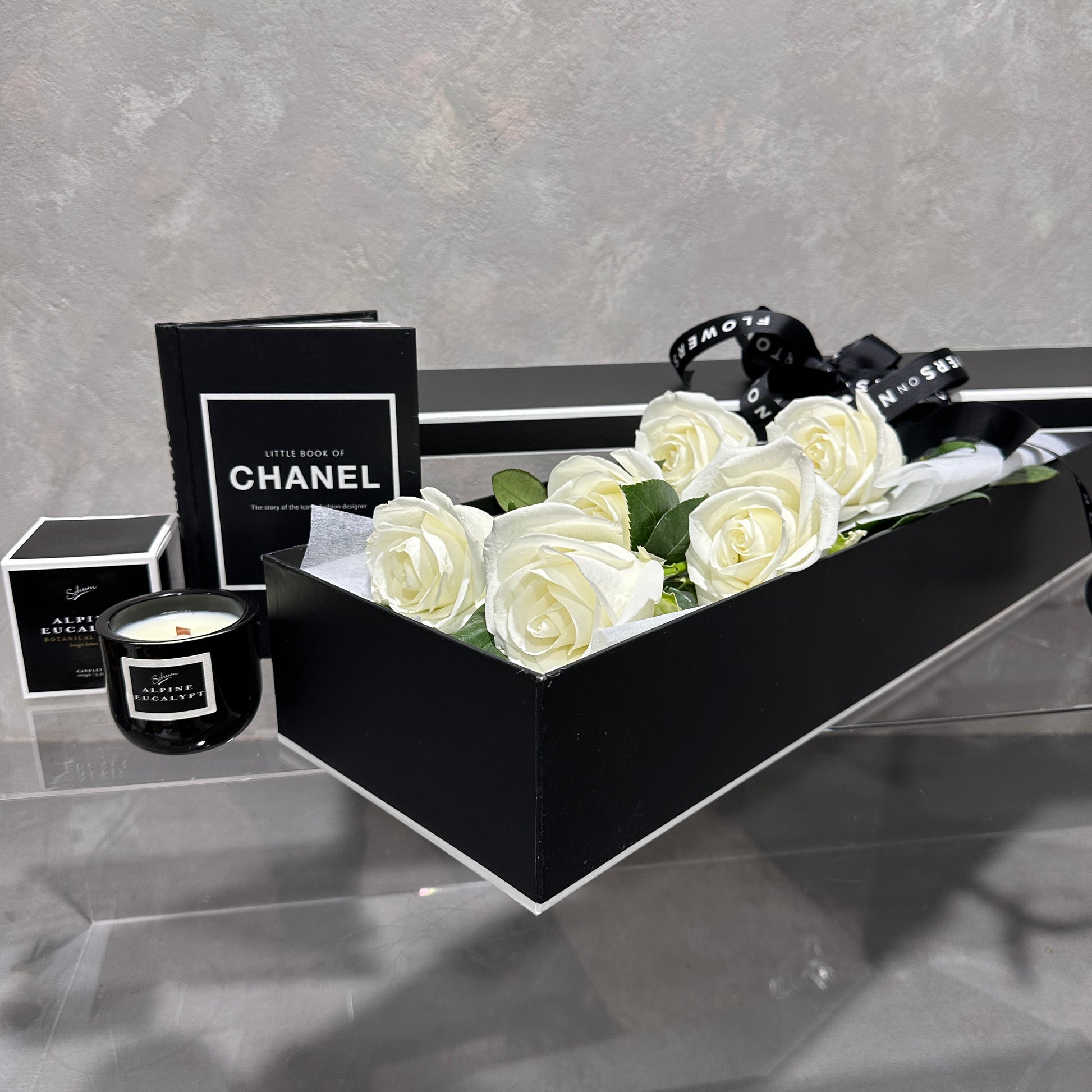 A FlowersonNortonSt Rose Gift Box, featuring a sophisticated black exterior and filled with pristine white roses, is elegantly displayed on a clear surface. Nearby, you can find a stylish black candle in a jar and the chic "Little Book of Chanel" in black. A striking black ribbon with white text adds to this memorable gift setting.