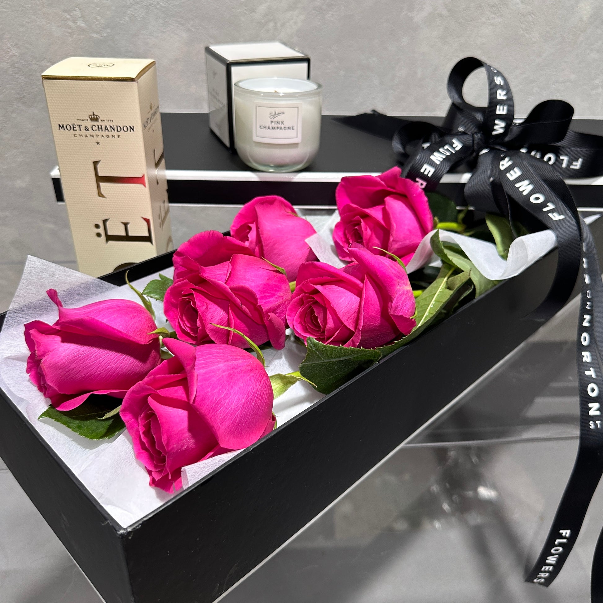 FlowersonNortonSt's The Rose Gift Box showcases vivid pink roses beautifully arranged in a sleek black box adorned with a matching ribbon. This exquisite presentation is complemented by Moët & Chandon packaging and a white candle, crafting an unforgettable and sophisticated gift.