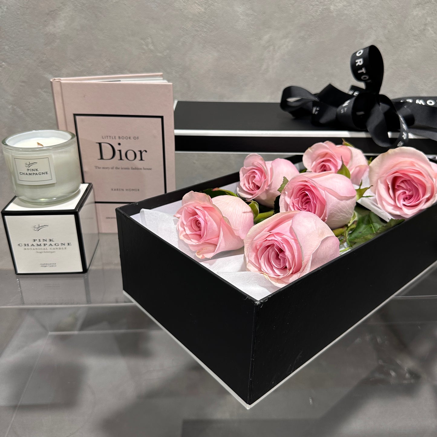 A black "The Rose Gift Box" by FlowersonNortonSt, containing pink roses, is elegantly displayed on a glass surface next to the "Little Book of Dior." A lit "Pink Champagne" scented candle with its matching box enhances the presentation's sophistication. A black ribbon is partially visible in the background, creating a memorable gift display.