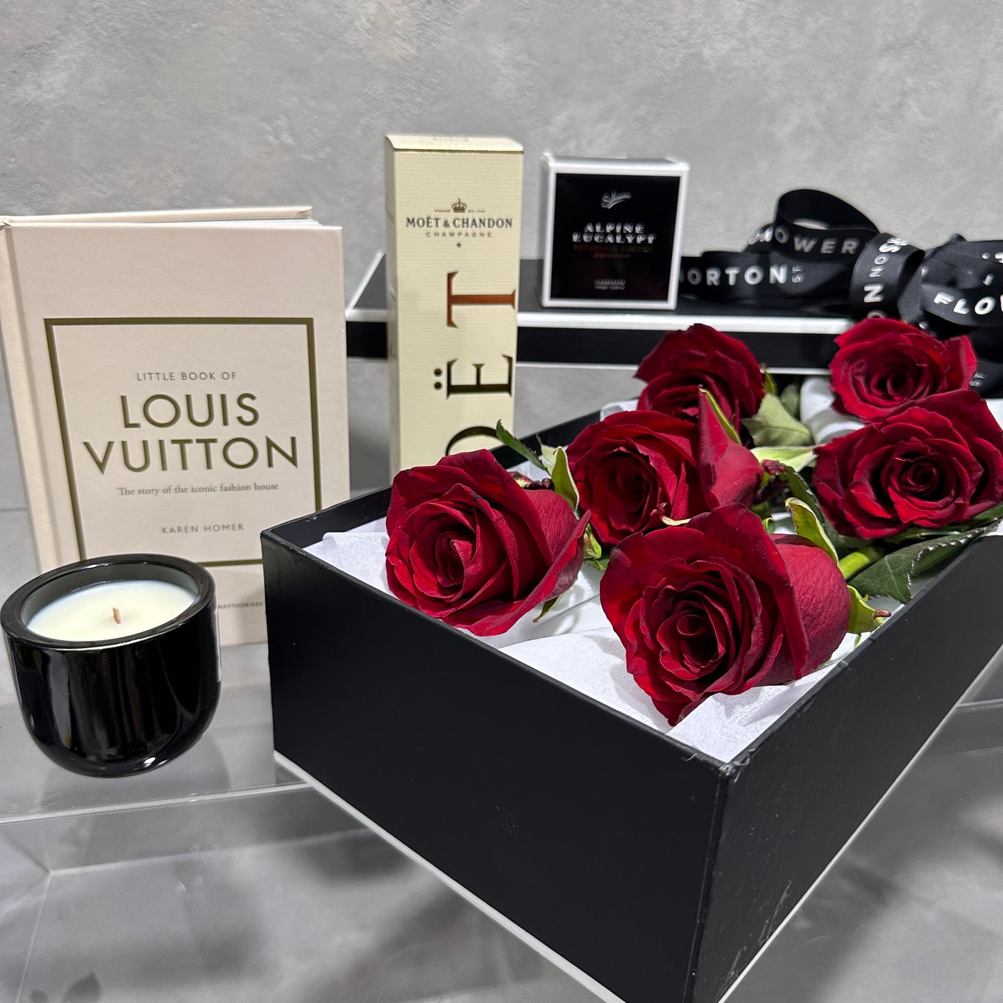 A memorable gift display featuring FlowersonNortonSt's The Rose Gift Box with red roses, a candle, the "Little Book of Louis Vuitton," a Moët & Chandon box, and other assorted packaged delights, all elegantly set on a glass table.