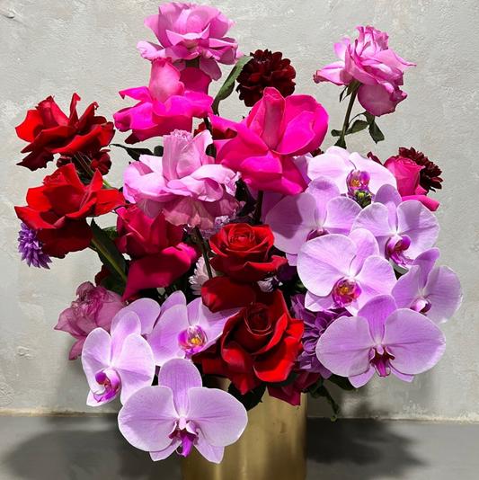 The FlowersonNortonSt Passion Pop Purple Bouquet in a gold pot features deep red roses, magenta flipped roses, and light purple phalaenopsis orchids, creating a lush and vibrant display against a neutral background.