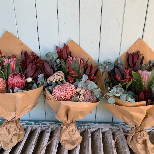 The "Little Native Bunch" from Flowers on Norton St features three rustic bouquets wrapped in brown paper, showcasing pink proteas, red and pink flowers, and eucalyptus leaves, creating a statement bouquet with seasonal blooms. These arrangements are beautifully displayed against a light blue wooden background to celebrate the beauty of native flora.